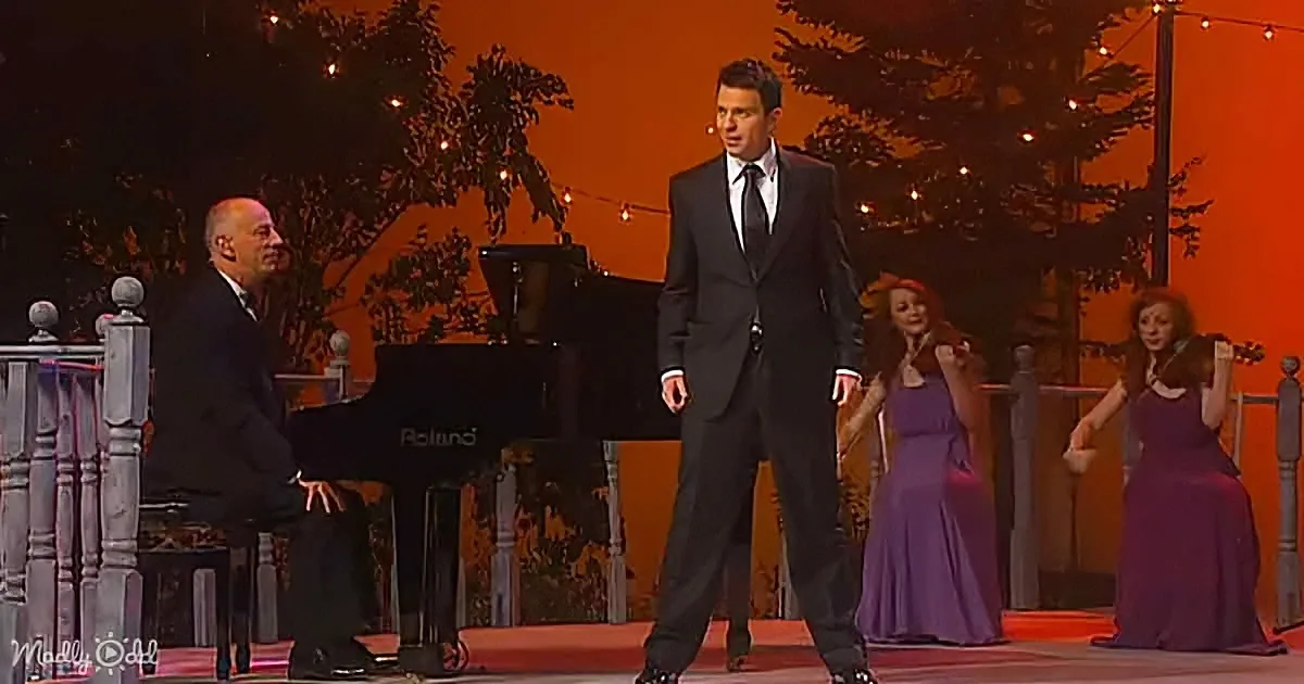 A man in a suit dances on stage with musicians and a violinist in purple dresses.