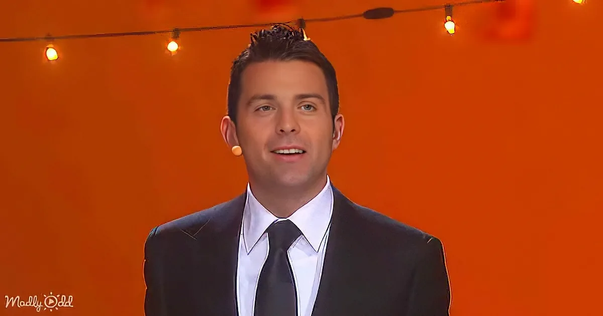 A man in a suit and tie stands against an orange backdrop with string lights.