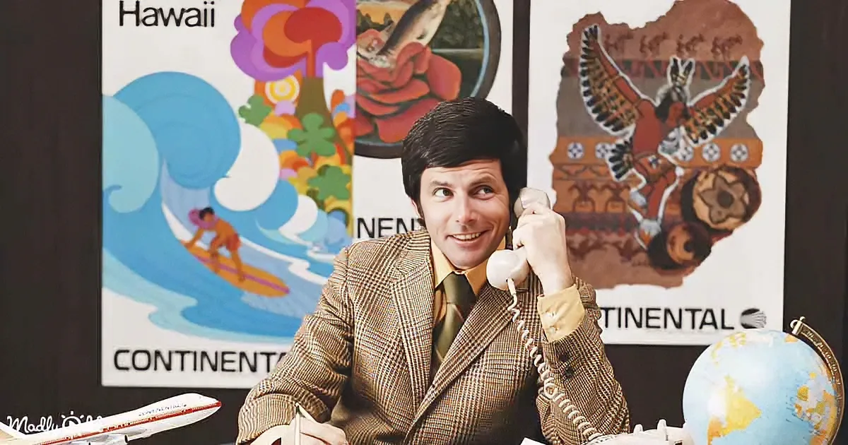 A man in a brown suit smiles while talking on a phone, surrounded by travel posters and a globe.