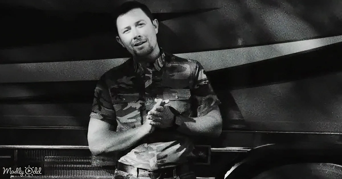 A man in a camouflage shirt sits with his hands clasped, leaning against a piano, in a black-and-white image.