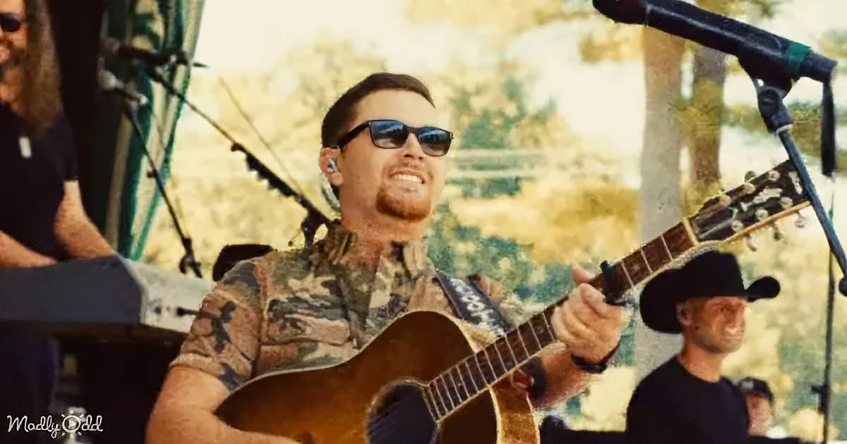A man in sunglasses plays an acoustic guitar on stage, surrounded by microphones and other musicians.