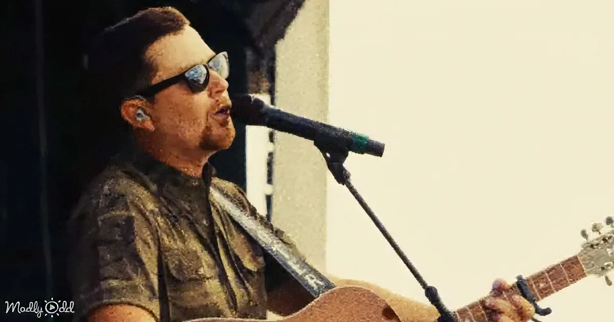 A man in sunglasses sings into a microphone while playing an acoustic guitar.