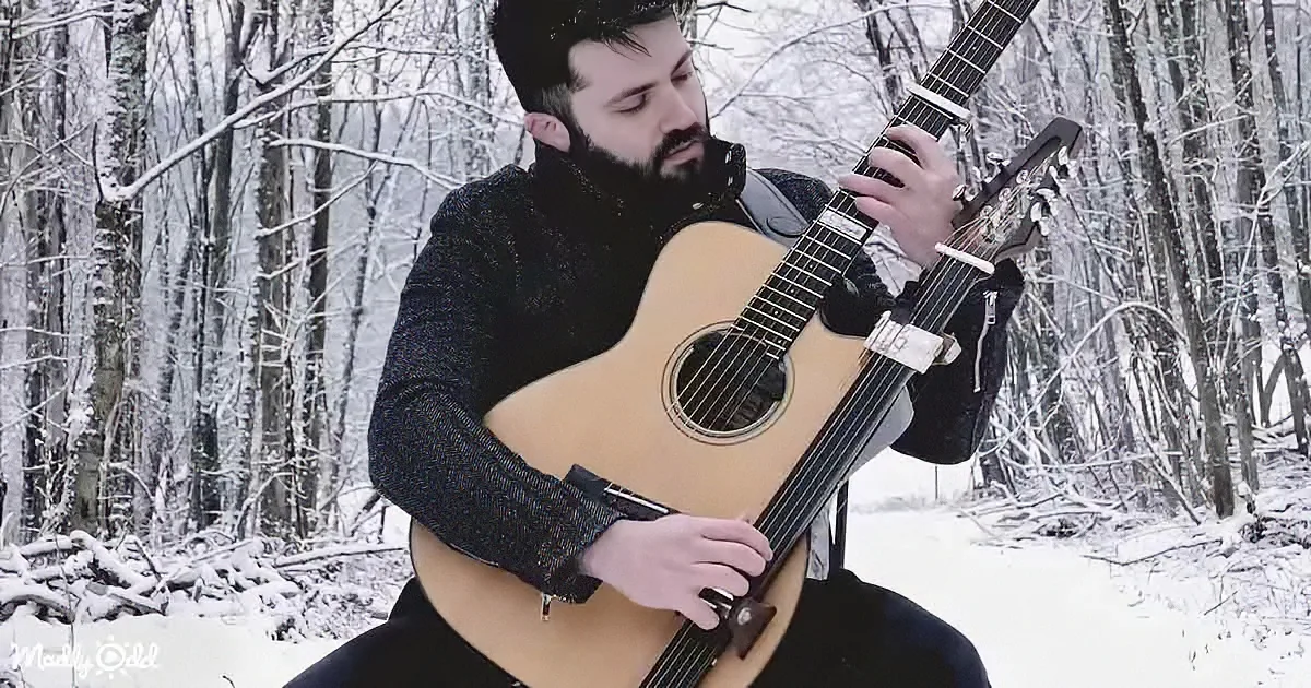 A man in a snowy forest plays an acoustic guitar, wearing a dark sweater and a scarf.