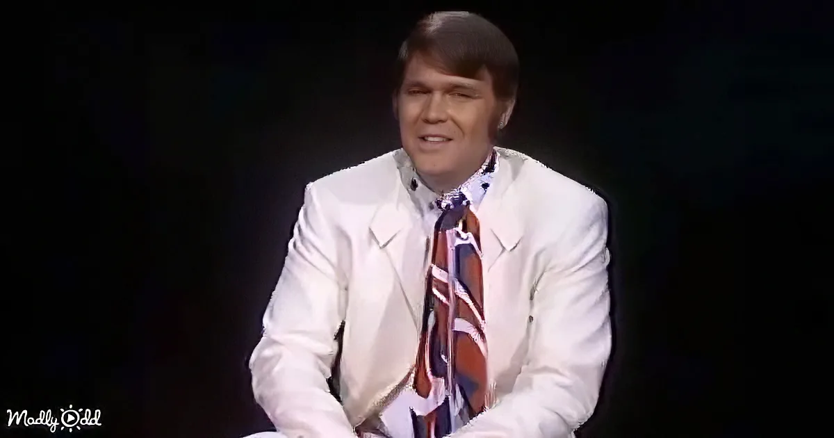 A man in a white suit and patterned tie sits against a dark background, smiling.