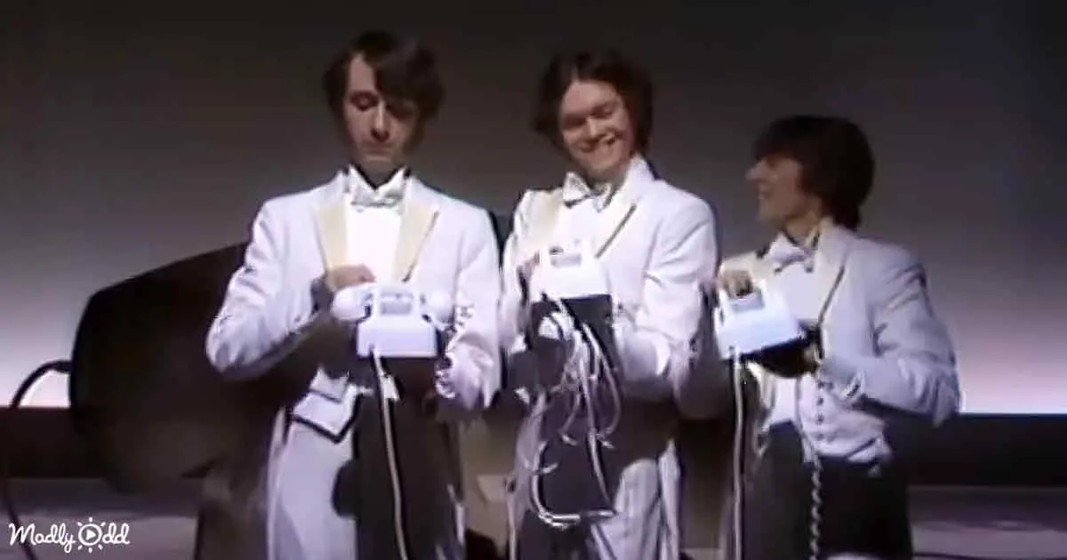 Three men in white suits hold vintage gaming controllers, smiling and posing together.