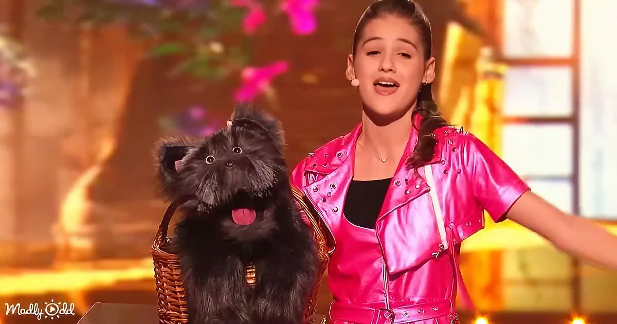 A girl in a pink outfit sings on stage with a black dog in a basket.