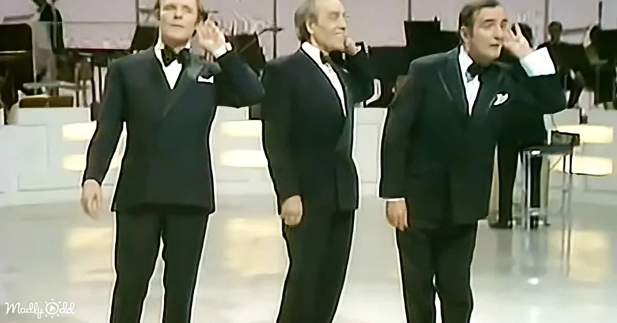 Three men in black suits and bow ties perform on stage with a band in the background.