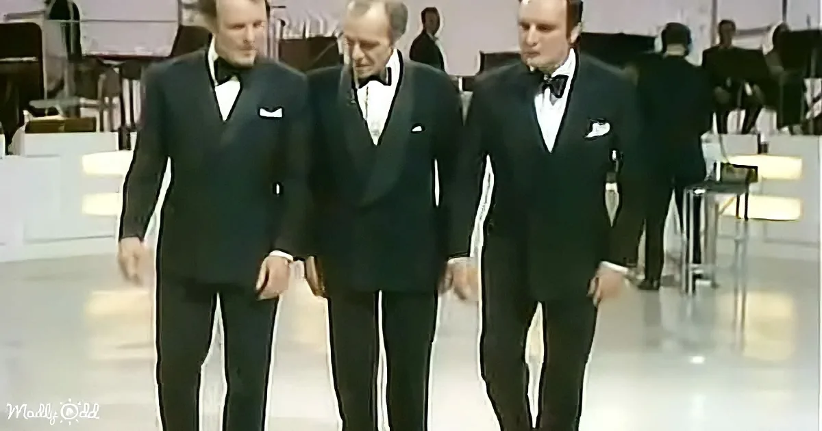 Three men in black tuxedos walk together in a formal setting, possibly a performance or event.
