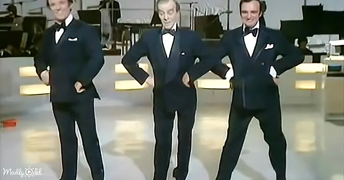 Three men in tuxedos pose confidently on a stage with an orchestra in the background.