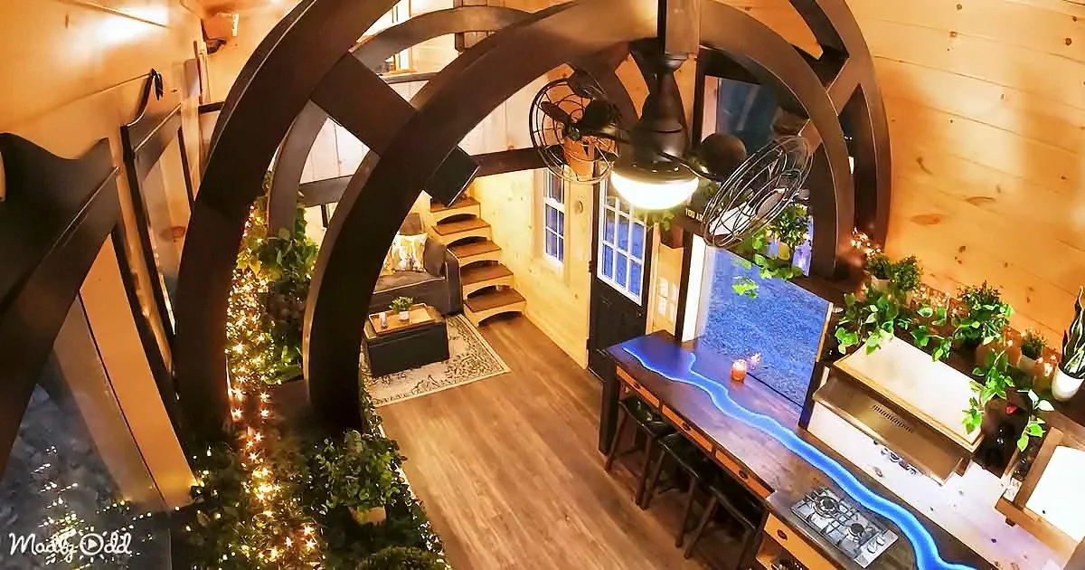 A cozy, rustic interior with a large wheel structure, wooden floors, plants, and a blue-lit table.