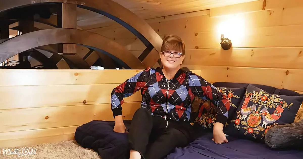 A woman in a plaid hoodie sits on a bed in a cozy, wooden cabin with a spiral staircase in the background.