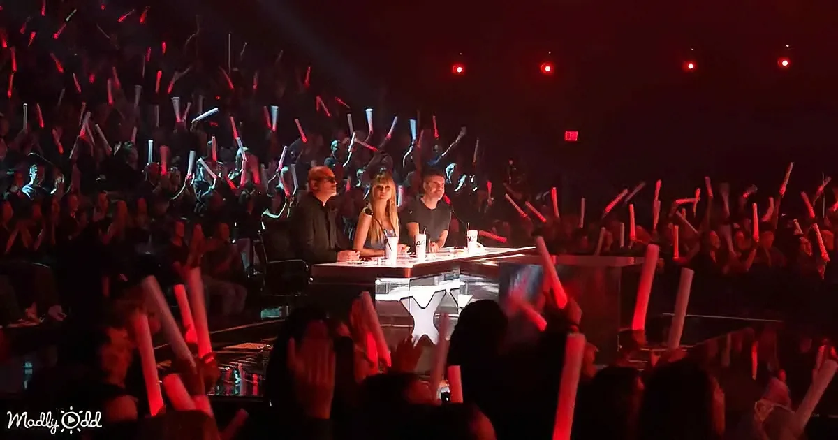 Judges on stage with audience raising light sticks in a dark venue.