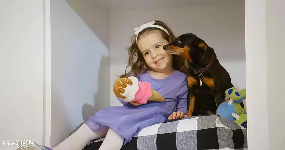 A young girl in a purple dress sits on a bed, holding a doll, with a black and brown dog next to her.