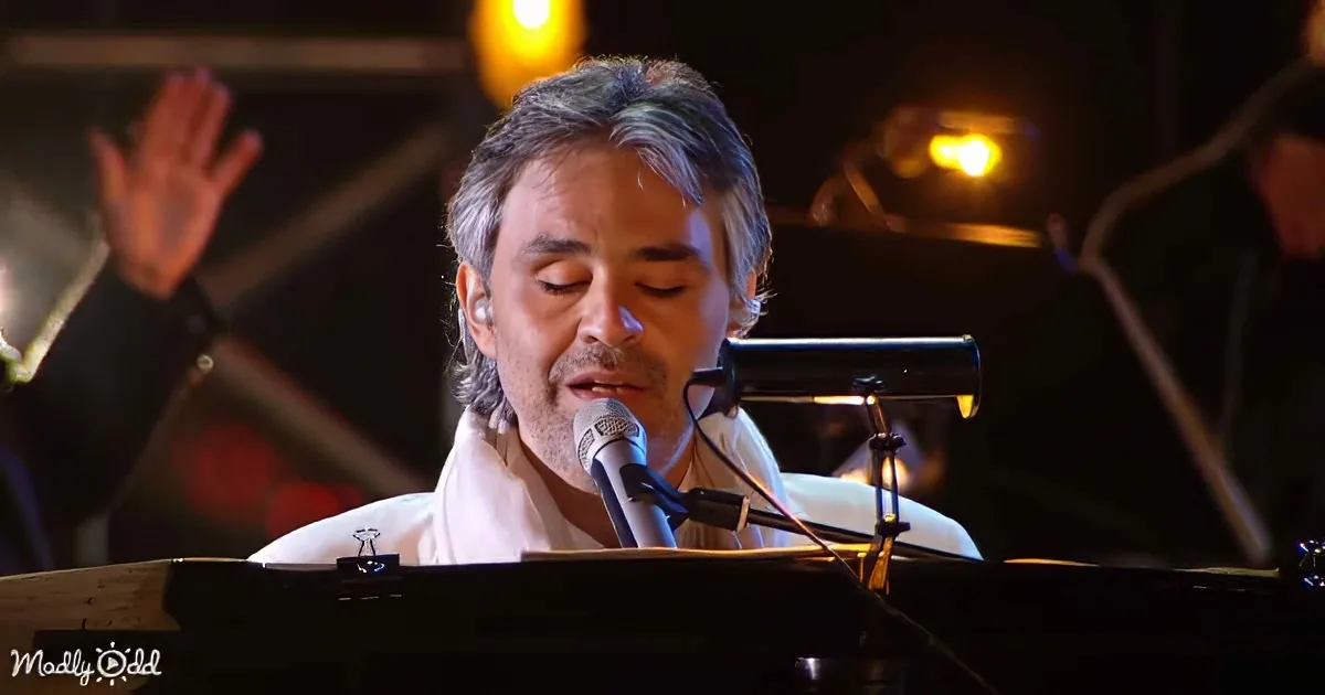 A man with gray hair and beard sings into a microphone while playing a keyboard, with a blurred background of lights and...