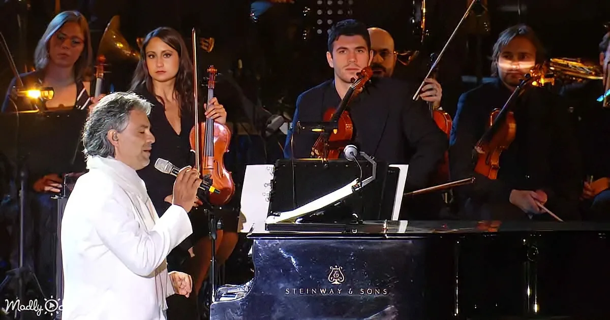 A man in white sings into a microphone beside a Steinway & Sons piano, with an orchestra behind him.