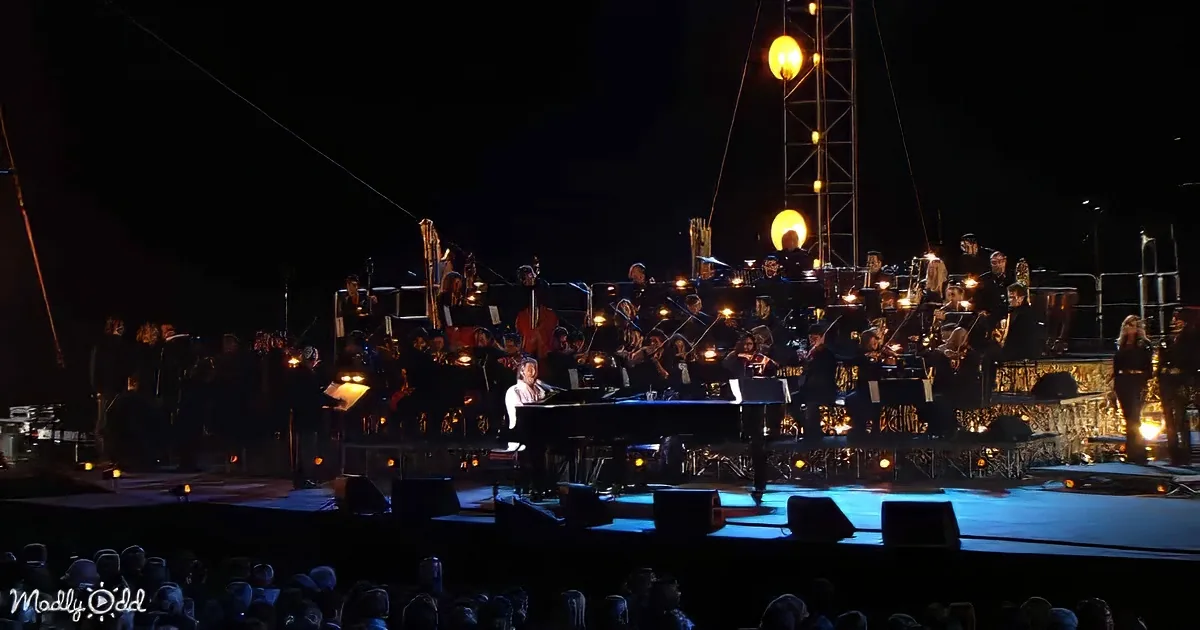 A pianist performs on stage with an orchestra, illuminated by stage lights, in front of an audience.