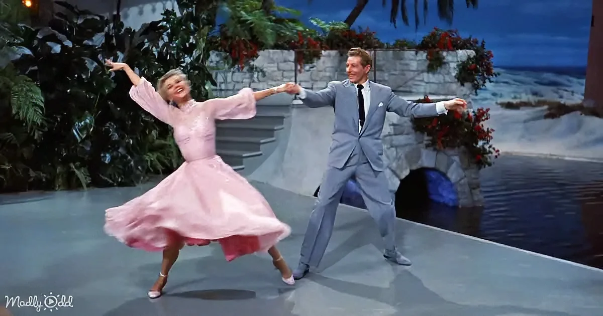 A man and woman in vintage attire dance joyfully on a stage with a tropical backdrop.