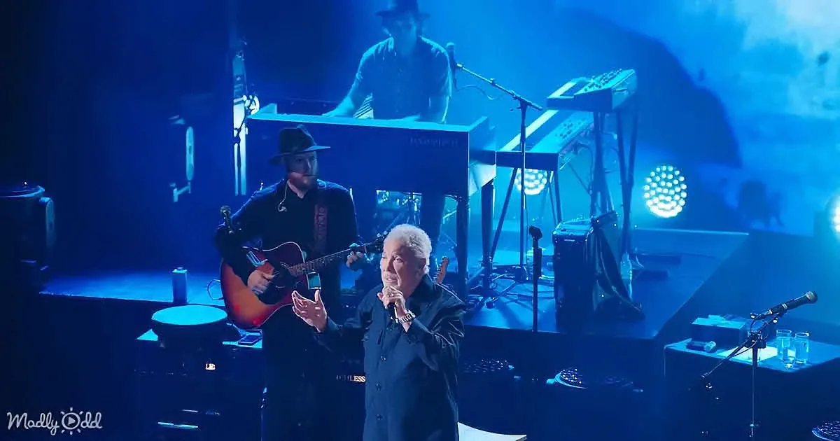 A man in a suit sings into a microphone on stage, accompanied by musicians and blue lighting.