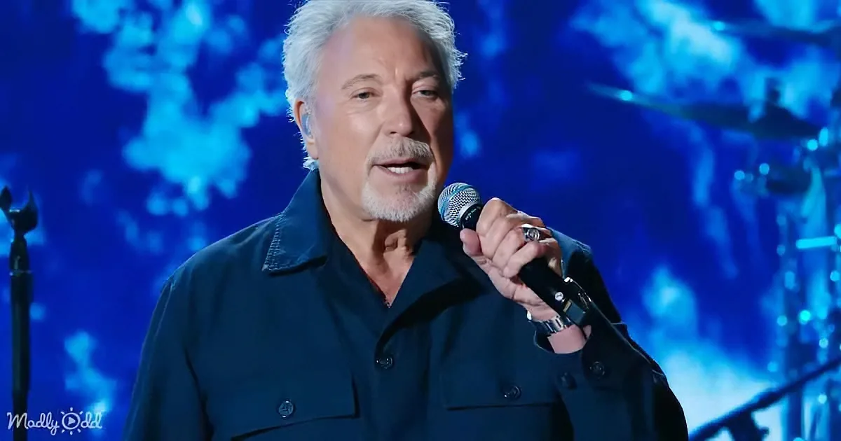 A man with white hair and a beard sings into a microphone on stage, wearing a dark jacket.