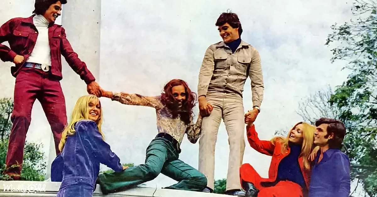 Five people in 1970s fashion hold hands, forming a circle on a white surface outdoors.