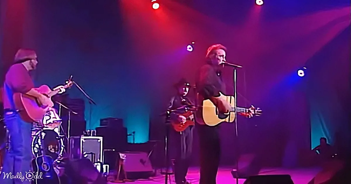 A man with long hair and a hat plays guitar and sings on stage under colorful lights.