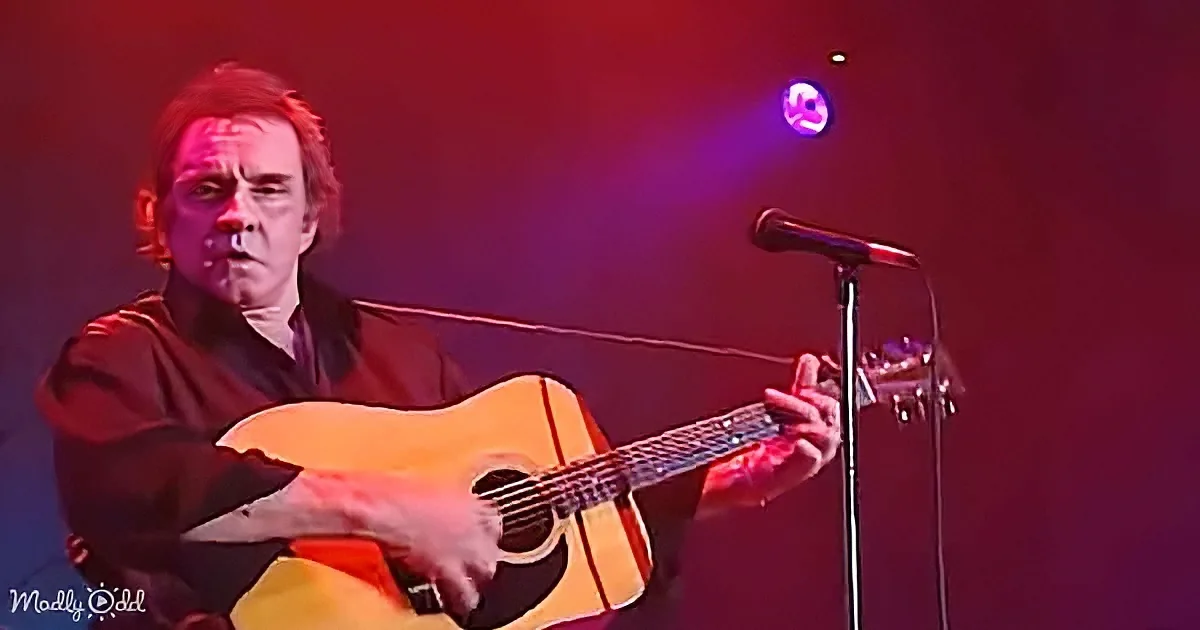 A man plays an acoustic guitar on stage with a microphone and red lighting.