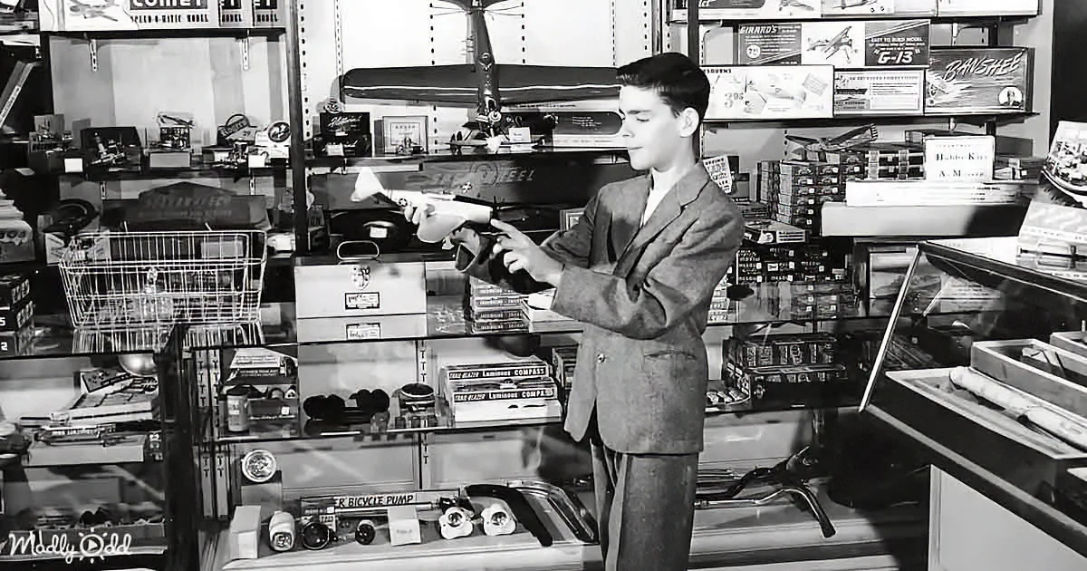 A boy in a suit holds a toy airplane in a store filled with model kits and other items.
