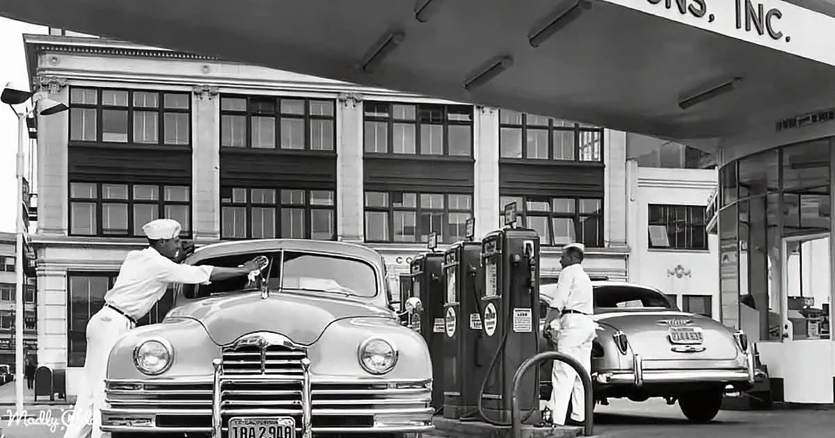 A man in a white uniform fills a vintage car's gas tank at a gas station.