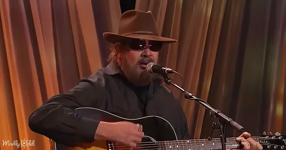 A man in a hat and sunglasses sings into a microphone while playing an acoustic guitar.