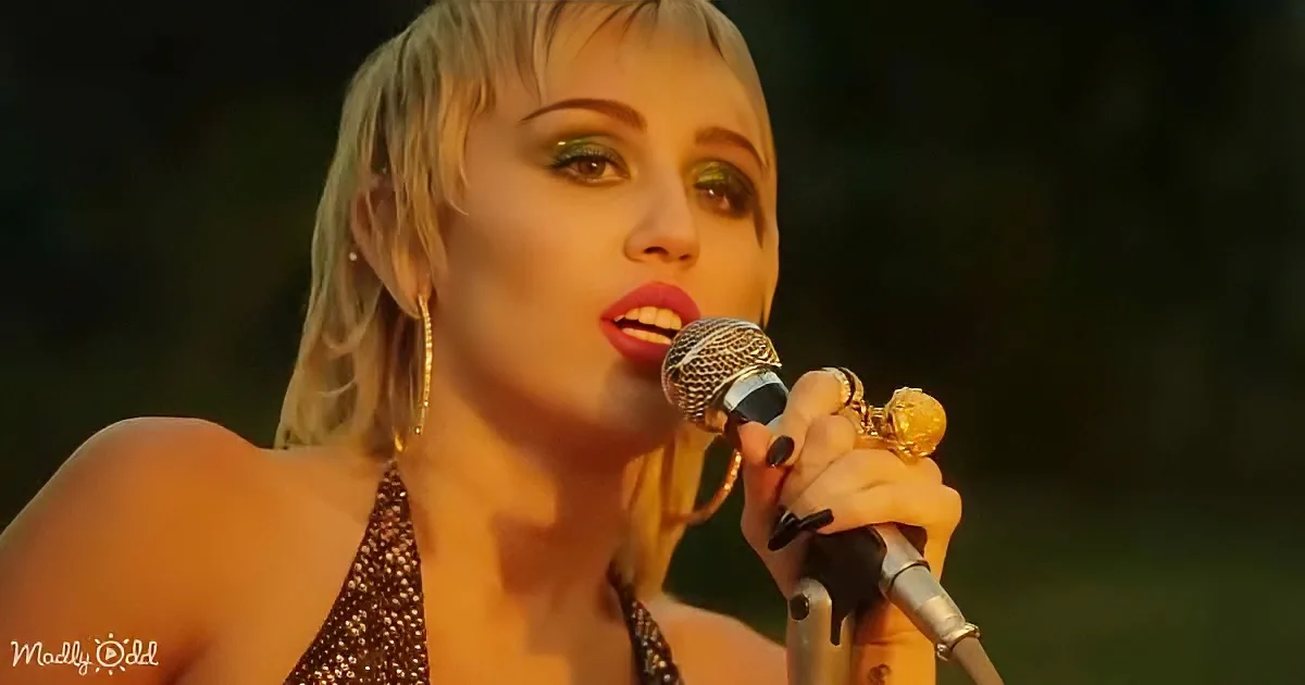 A woman with short blonde hair and red lipstick sings into a microphone, wearing a sparkly top and gold hoop earrings.