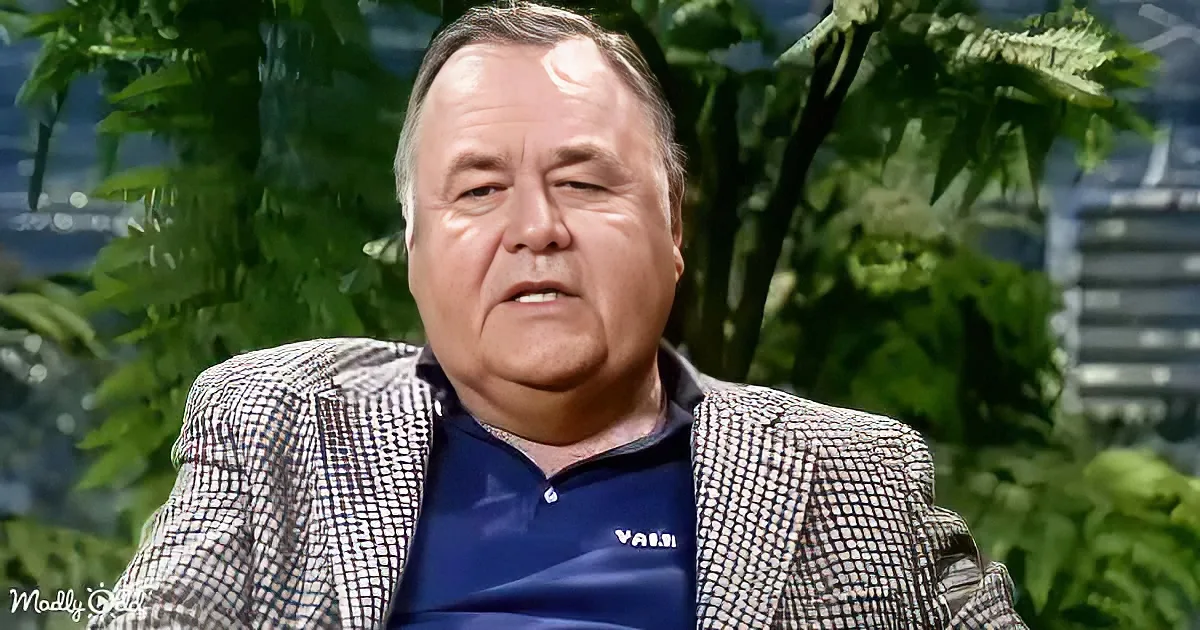 A man in a checkered blazer and blue shirt sits against a green leafy background.