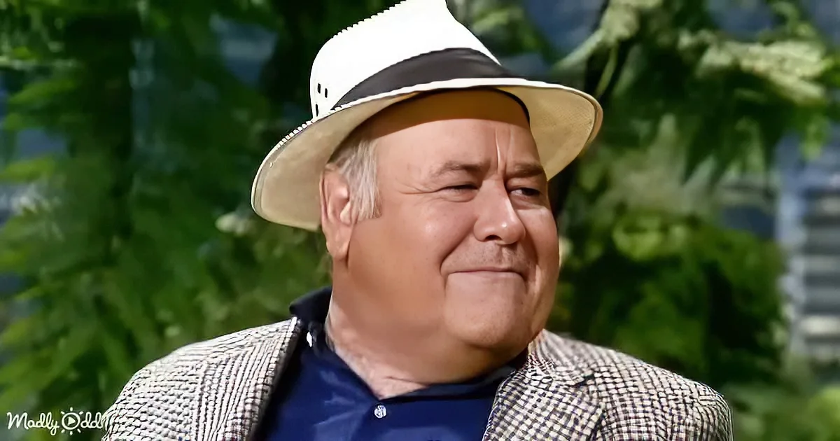 A man wearing a white hat and a checkered jacket smiles with a green, leafy background.