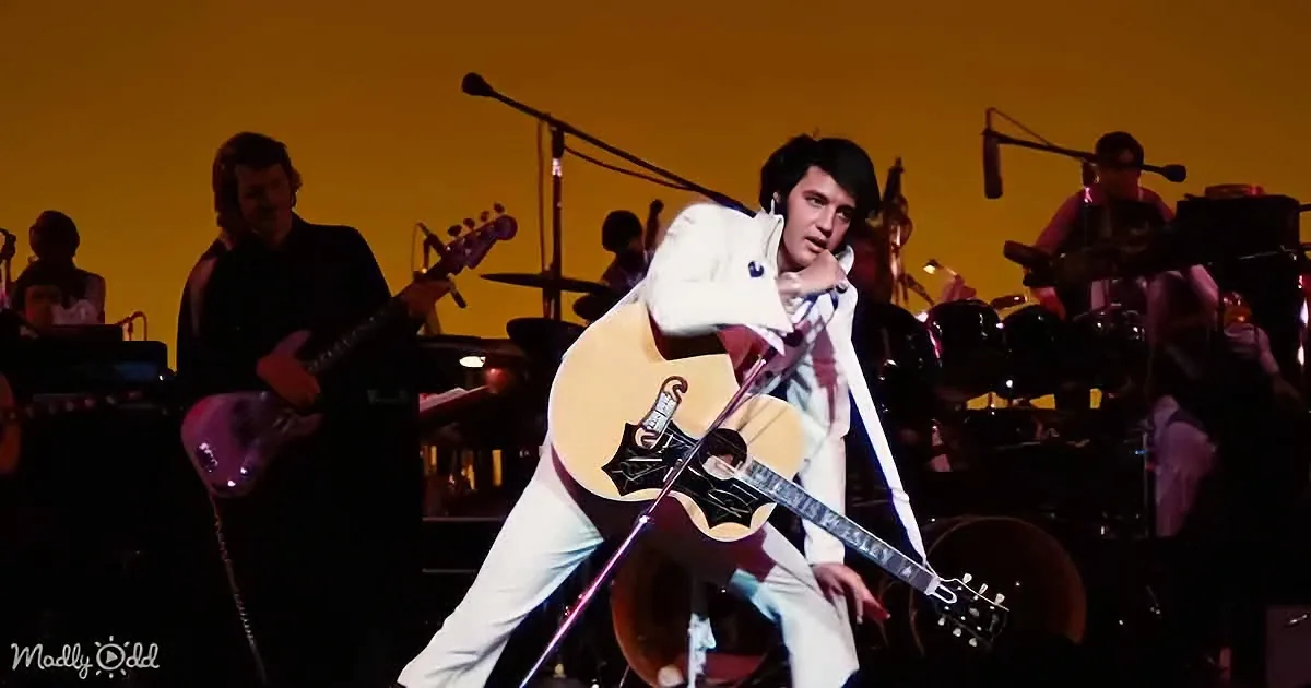 A man in white plays a guitar on stage with a band behind him.