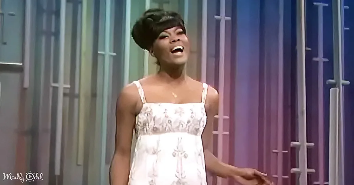 A woman in a white dress with a floral pattern is singing on stage with a colorful background.