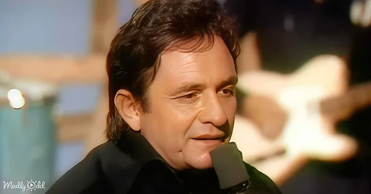 A man with dark hair and a black shirt is speaking into a microphone, with a blurred background and a light source visible.