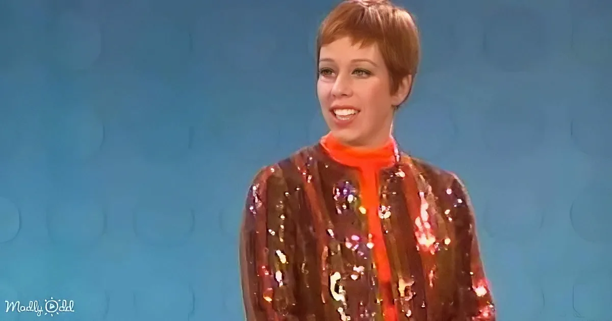 A person with short hair, wearing a shiny, patterned jacket over an orange turtleneck, stands against a blue background.