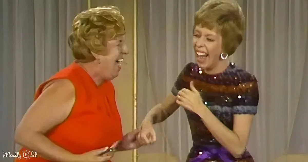 Two women in vintage attire are laughing and interacting on a stage, with one in an orange dress and the other in a...
