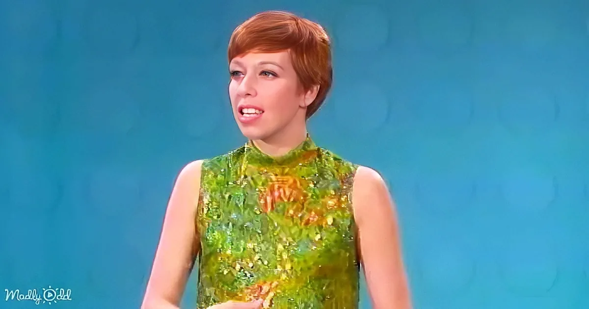 A woman with short red hair in a green sequined dress stands against a blue background.