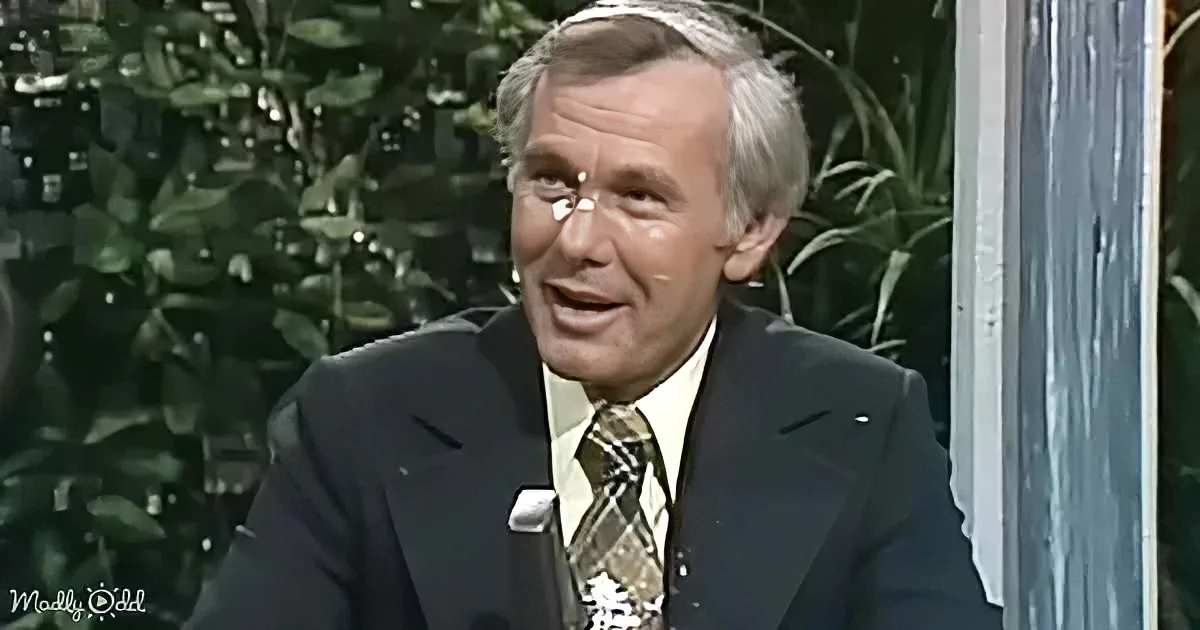 A man in a suit and tie, smiling, with a green leafy background.