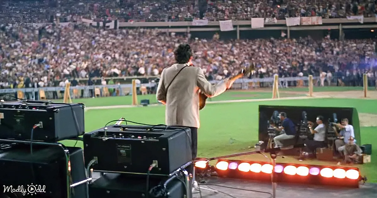 A man in a suit plays guitar on a baseball field stage, with a large crowd in the background.