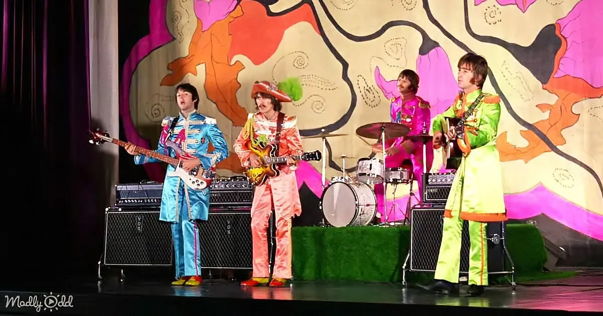 Four musicians in colorful outfits perform on stage with a psychedelic backdrop.