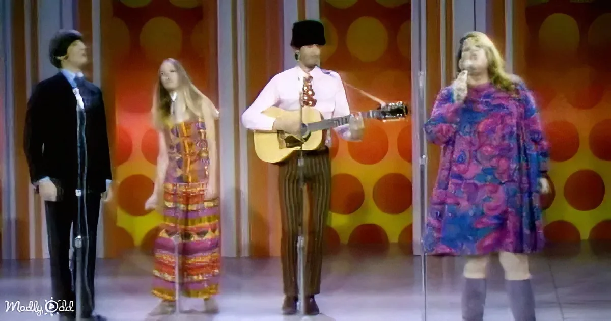 A man in a hat plays guitar while three others in colorful outfits stand nearby on a stage with a polka-dot background.