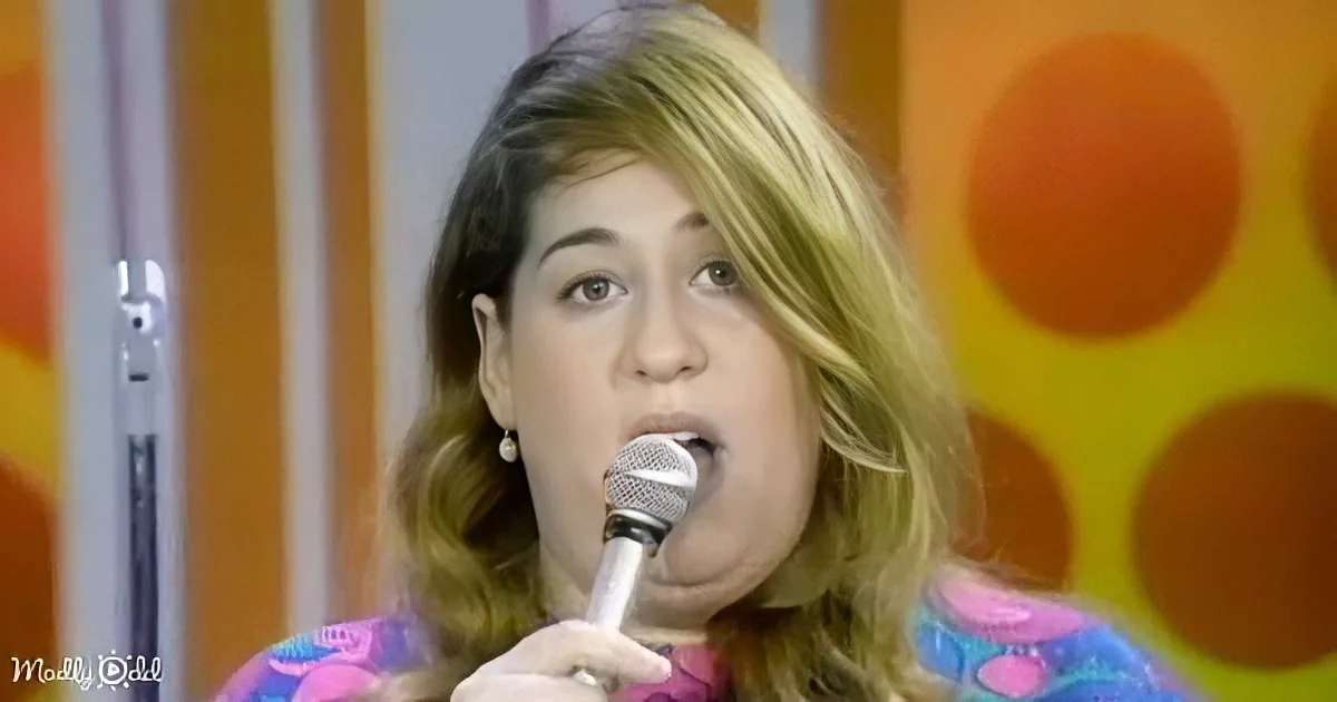 A woman with shoulder-length hair sings into a microphone, wearing a colorful top, against a vibrant orange and yellow...