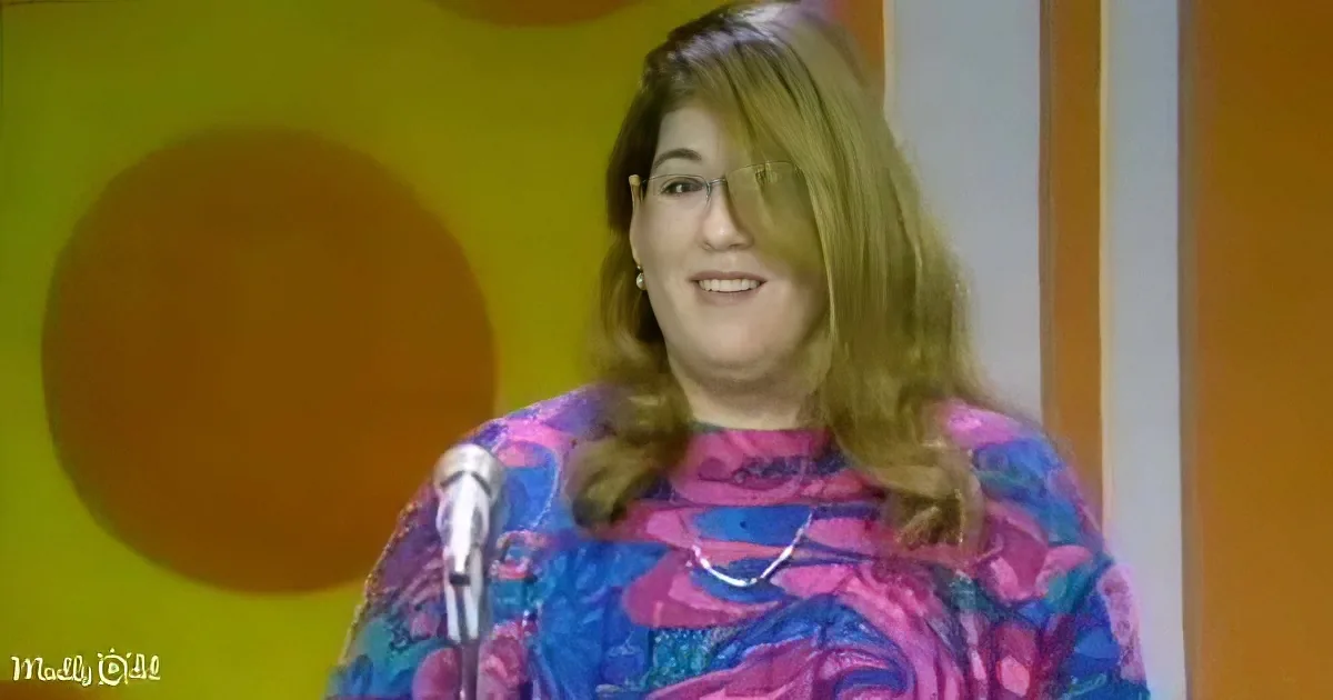 A woman with shoulder-length hair, wearing a colorful patterned top, smiles near a microphone against a yellow and orange...