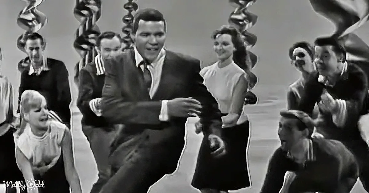 A man in a suit dances energetically in front of a group of people, all smiling and watching.