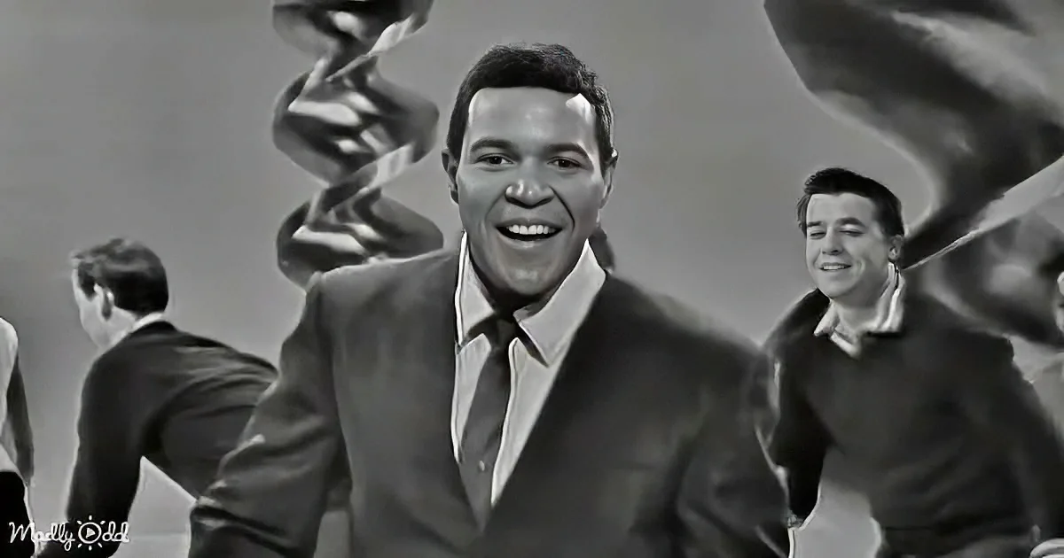 A man in a suit smiles brightly, surrounded by others in a lively setting.