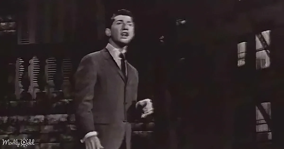 A man in a suit and tie stands on a stage, singing with his eyes closed.