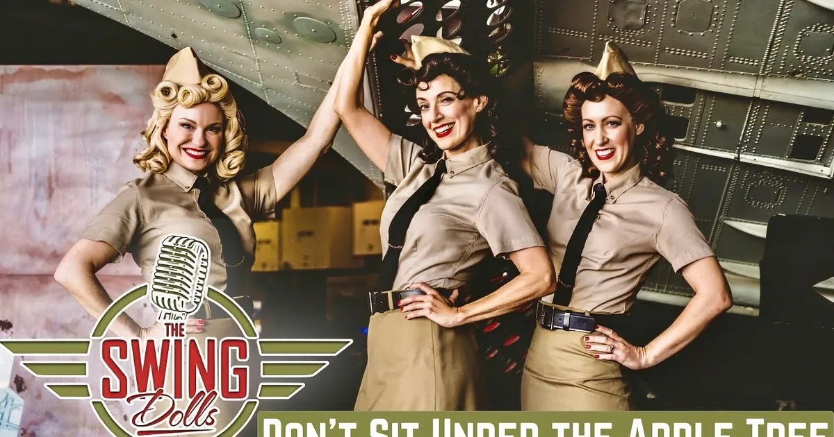 Three women in uniform-style outfits sing on stage with a band in the background.