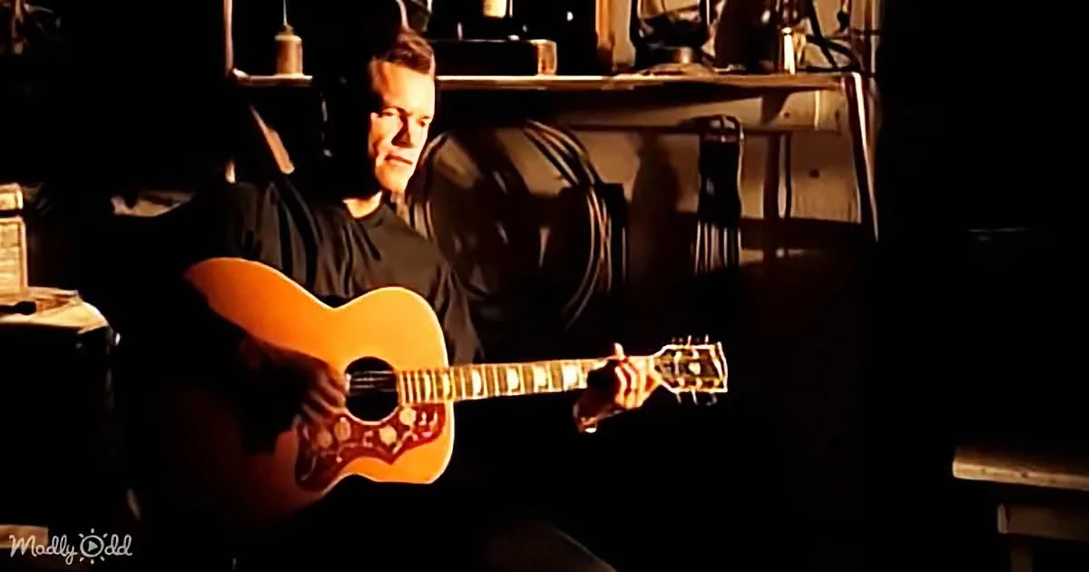 A man plays an acoustic guitar in a dimly lit room with a bottle of beer nearby.