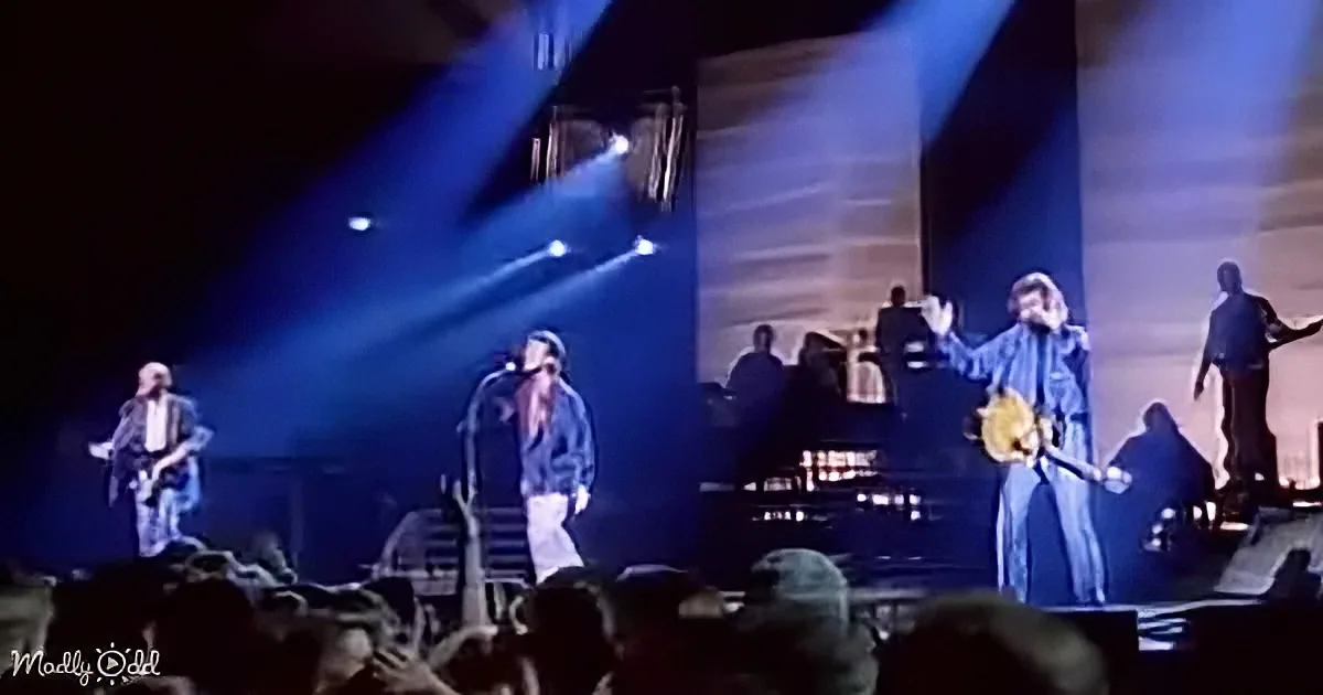 A band performs on stage with blue lighting, audience in foreground, and musicians playing instruments.